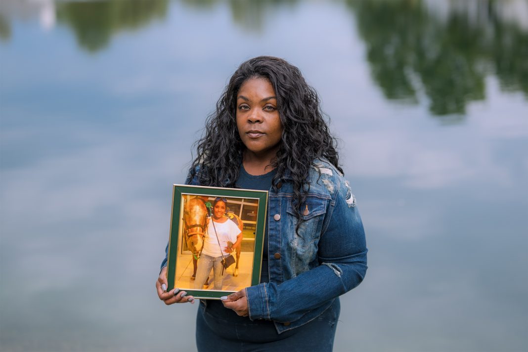 A photo of a Black woman standing by water. She holds a framed photo of her deceased mother.