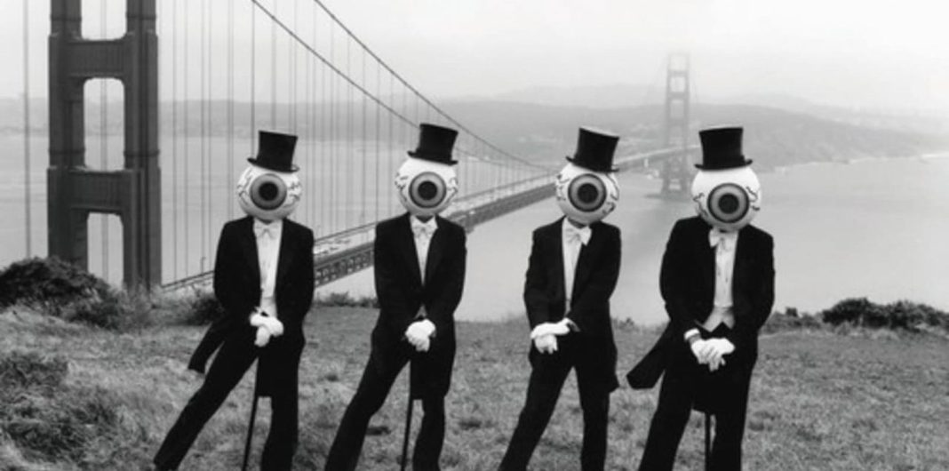 The Residents wearing eyeball head masks and tuxedos seated in front of the Golden Gate Bridge in San Francisco