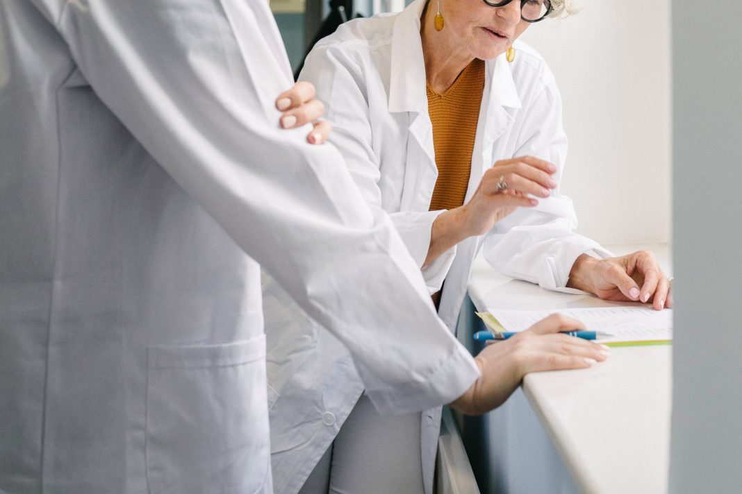 An older doctor consults with a younger doctor as they both review forms on a clipboard.